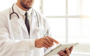 A imagem mostra um médico, de pele branca, utilizando um tablet. Ele usa um jaleco branco e um estetoscópio preto com prata sobreposto em seus ombros. Por baixo do jaleco, é possível visualizar uma camisa clara e uma gravata preta. Ele está voltado para o lado direito da imagem, em pé, segurando um tablet branco com sua mão esquerda, enquanto seu dedo indicador direto toca a tela.