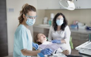 A imagem retrata um consultório odontológico. Em primeiro plano está uma mulher branca e loira. Seus cabelos estão presos, ela veste um jaleco azul, luvas, máscara e óculos de proteção. Ela manuseia utensílios de dentista. Em segundo plano e desfocadas, estão duas mulheres brancas. Uma delas é loira e está deitada em uma maca. Ao seu lado está sentada uma mulher com cabelos escuros e que veste óculos de proteção, máscara e luvas. Ela coloca um sugador odontológico na boca da paciente deitada.
