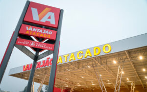 Na imagem, vemos a frente do mercado. Em primeiro plano, uma estrutura com três placas onde, na superior, temos o VA (logo da empresa), com fundo vermelho e branco e laranja nas letras; na placa abaixo, está escrito Vantajão Atacado, em fundo vermelho e as palavras em branco e amarelo; na terceira placa, temos a informação que lê “uma empresa do Grupo Andreazza” e a logo do Andreazza (um A no meio de um contorno), tudo em branco. Ao fundo, temos a fachada do mercado, com Vantajão Atacado no display, em vermelho e amarelo sobre um fundo branco, e luzes acesas na parte interna da cobertura.