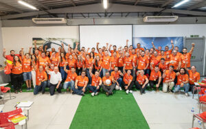 A imagem mostra um grupo de pessoas com camisetas laranjas. Eles estão posicionados ao centro da imagem, todos sorridentes e com as mãos erguidas para o alto. Em primeiro plano está um tapete verde, que se estende até o grupo de pessoas.