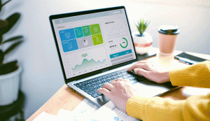 Na imagem há um notebook com informações a respeito da performance de uma empresa. Ele está em uma mesa de madeira, que possui alguns papeis, uma planta e um copo de café. A pessoa que está usando o notebook está usando um cardigan amarelo. Só aparecem seus braços. Na frente da mesa, há um vaso grande com uma planta.